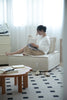 Woman sitting on a beige small sectional sofa using a laptop in a bright room with white curtains.