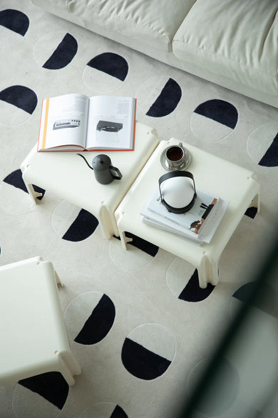 White small coffee table with black geometric patterns on a white rug with black shapes, featuring books and mugs.
