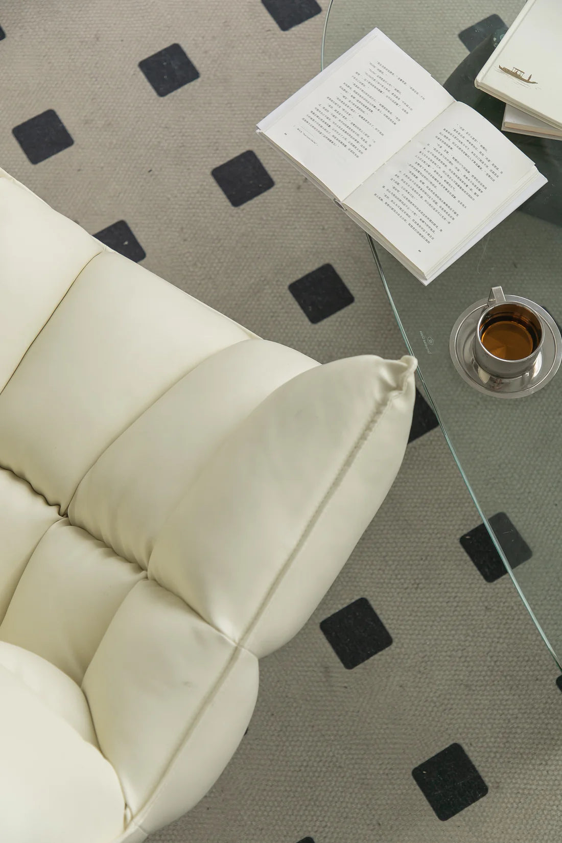 White lounge chair with a glass coffee table featuring an open book and a cup of coffee on a patterned rug.