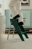 A person in white pants and black shoes steps onto a dark green metal step ladder with a silver frame in a modern room setting.