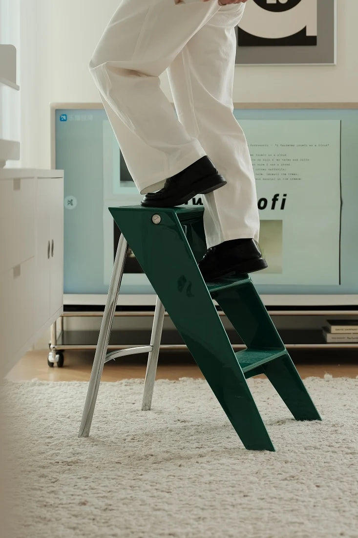 A person in white pants and black shoes steps onto a dark green metal step ladder with a silver frame in a modern room setting.