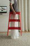 A person in a gray apron and white dress holds a bright red three-step ladder stool in a minimalist indoor setting with a beige carpet and cream curtains.