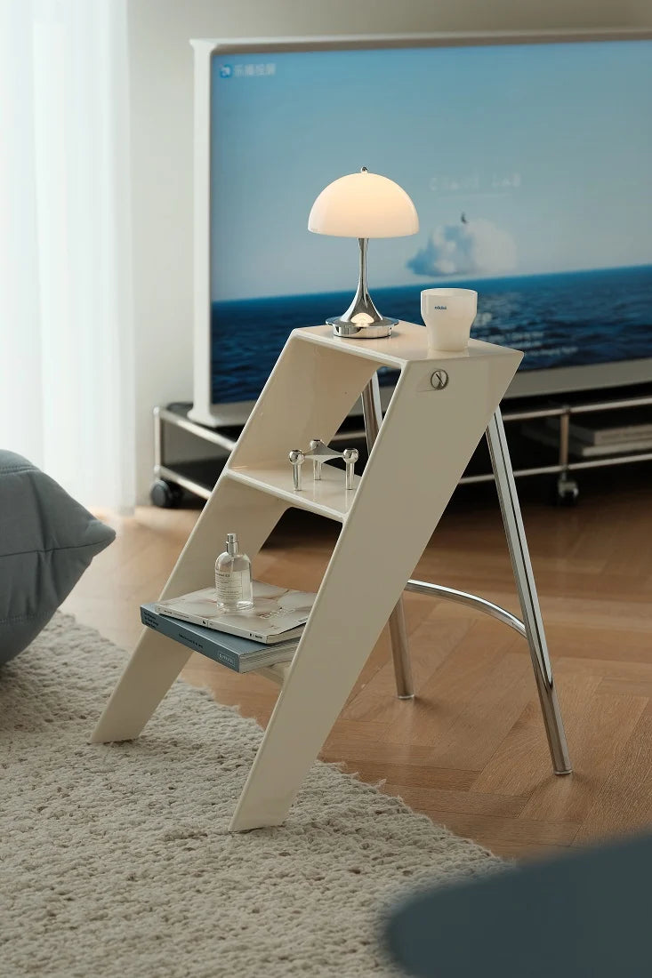 A modern A-frame side table in a light beige wood finish with chrome legs, styled in a living room setting with a lamp, cup, and tray on top, positioned next to a television.