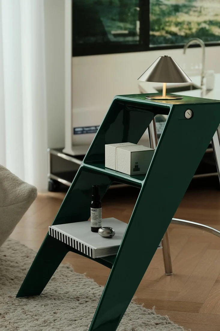 A modern dark green ladder-style side table in a living room setting, styled with a silver cone lamp, a white box, and a small bottle on its shelves.