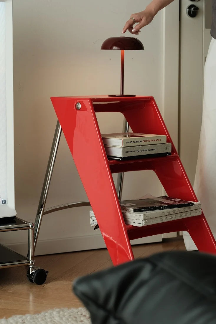 A red step ladder side table with three tiers, styled with books, next to a chrome rolling cart. A hand is shown turning on a dark red dome lamp placed on top of the table.