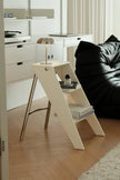 A modern cream-colored A-frame side table with a silver metal lamp and books, set in a minimalist living room next to a black leather sofa on a light wood floor.