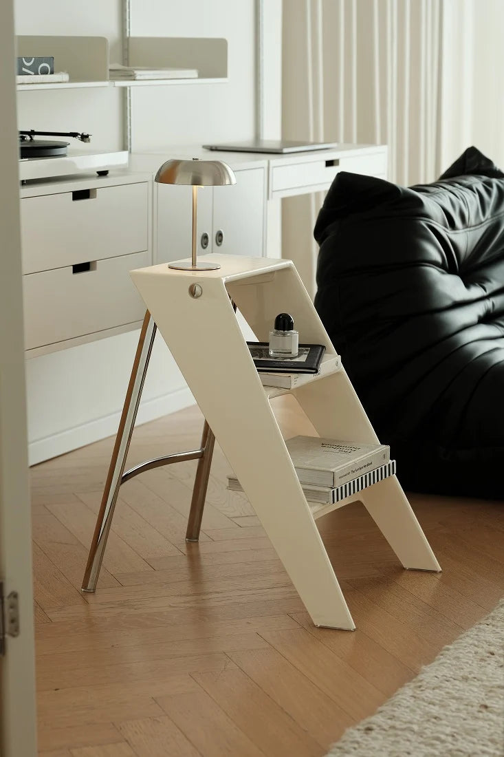 A modern cream-colored A-frame side table with a silver metal lamp and books, set in a minimalist living room next to a black leather sofa on a light wood floor.