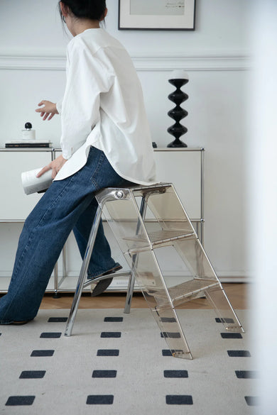 Lady using a transparent step ladder in a room with a white wall and black and white checkered floor.