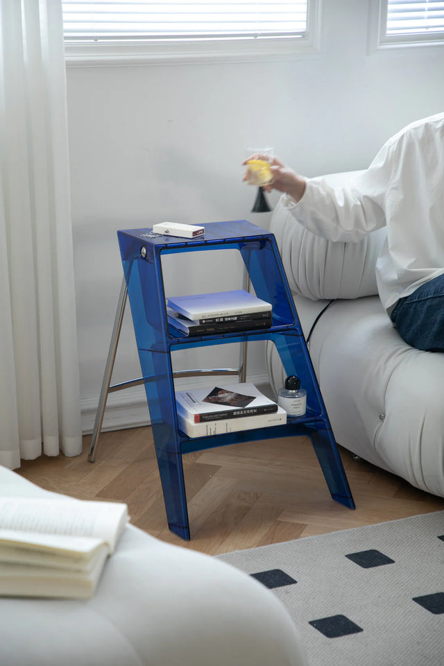 Blue step ladder shelf with books and a bottle on a wooden floor next to a white couch.