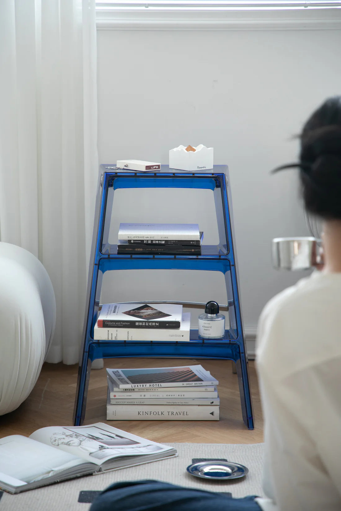 Lady sitting on a couch looking at a blue step ladder display shelf with books and a box on it.