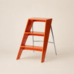 A glossy orange three-step ladder stool with a silver metal support frame, photographed on a clean, solid light beige background.