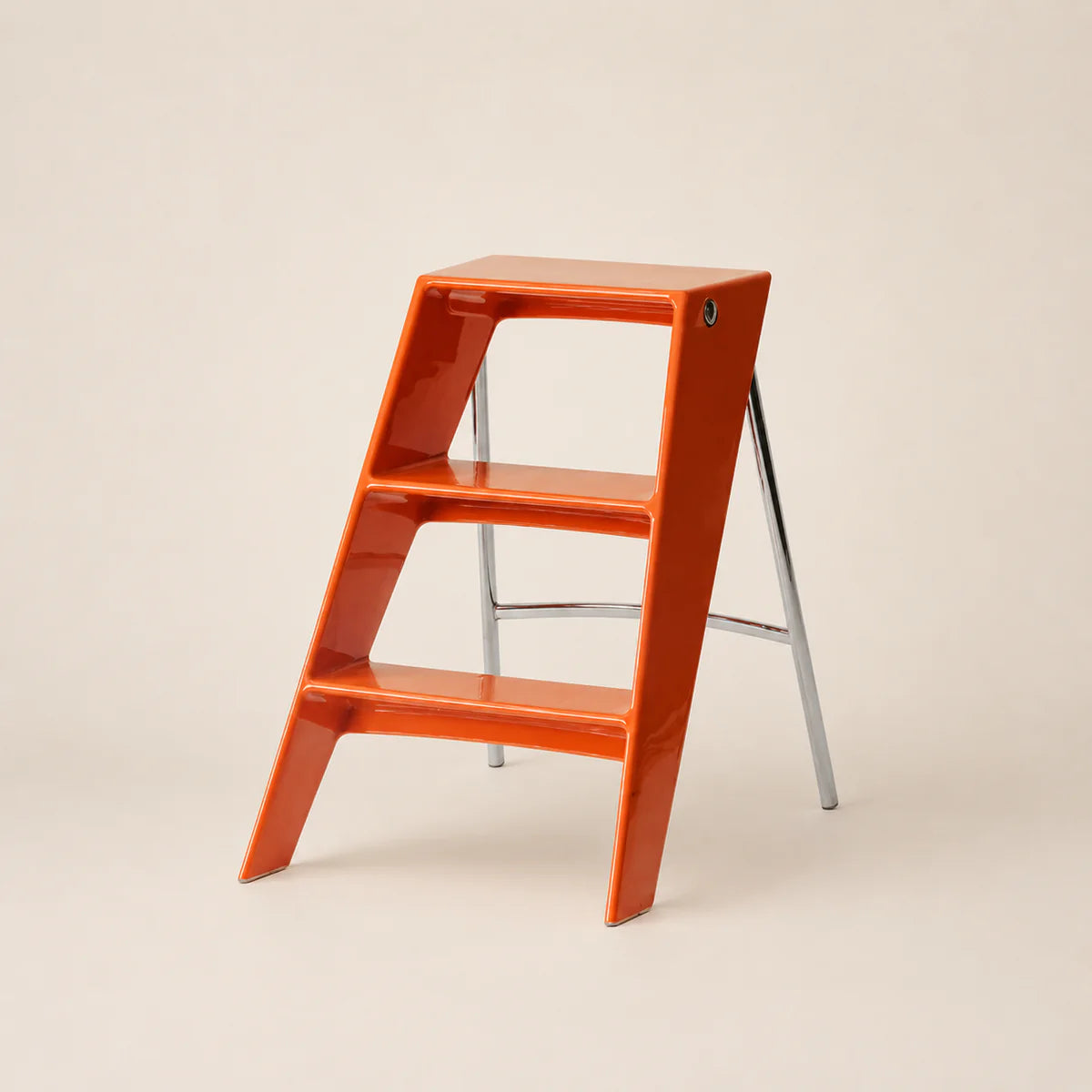 A glossy orange three-step ladder stool with a silver metal support frame, photographed on a clean, solid light beige background.