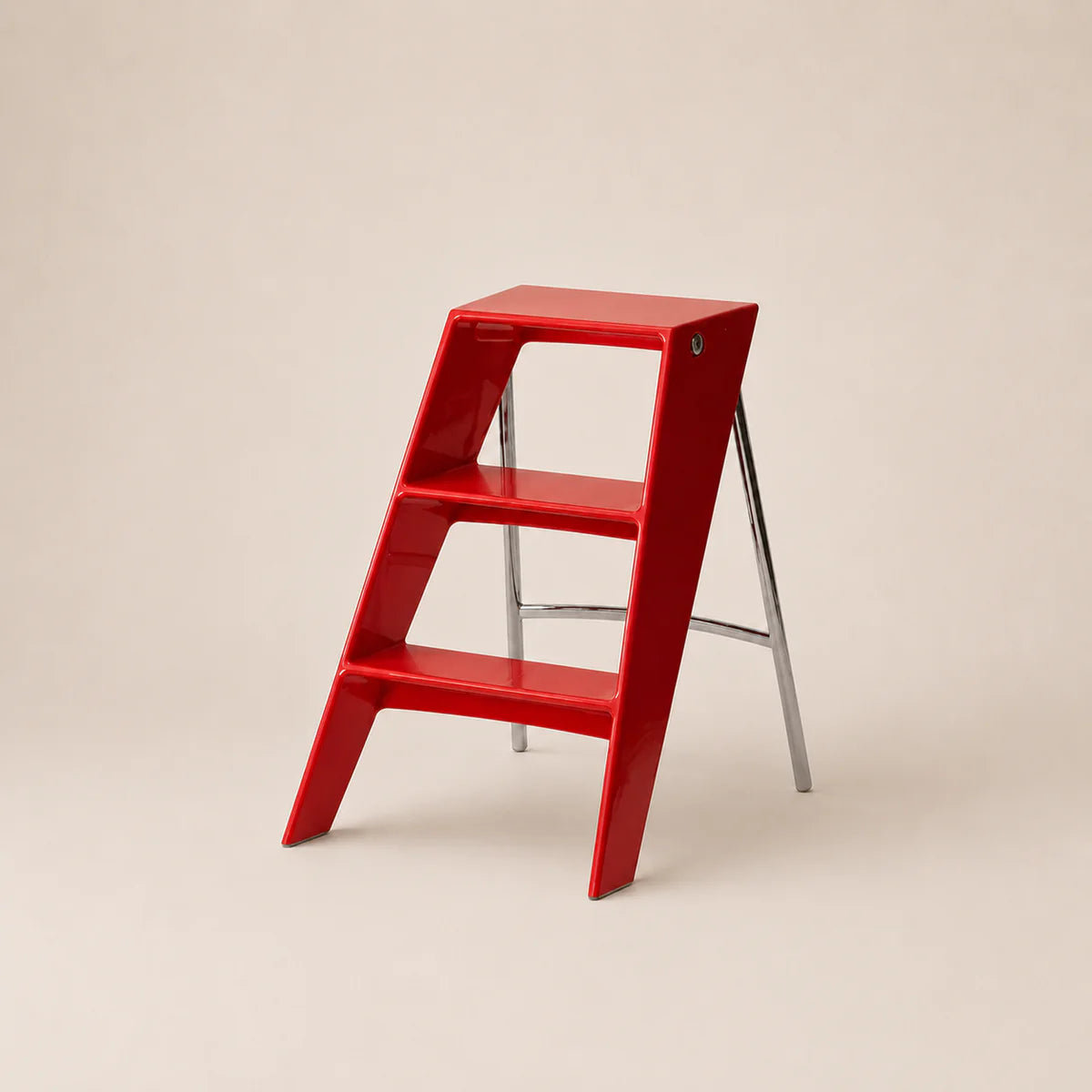 A ruby red three-step ladder stool with slim silver metal legs, photographed on a clean, solid beige background.