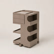 A modern, minimalist rolling storage cart in a warm brown color, featuring two drawers on the left, two open shelves, and three large circular cutout panels on the side, set against a clean, light beige background.