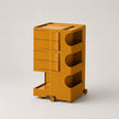 A mustard yellow storage cart with multiple drawers, open shelves, and small wheels, photographed on a clean light gray studio background.