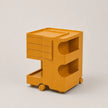 A golden yellow rolling storage cart with multiple drawers and open shelves, shown on a plain light gray studio background.