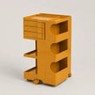 A bright yellow-orange rolling storage cart with a top tray, three drawers on the upper left, three open shelves on the right, and caster wheels, photographed on a light gray studio background.