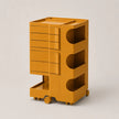 A mustard yellow rolling storage cart with multiple drawers and open shelves, photographed on a clean light gray studio background.