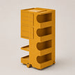 A mustard yellow plastic storage organizer with multiple drawers, open shelves, and caster wheels, shown on a neutral light gray background.