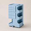 A light blue storage cart with multiple drawers, open shelves, and caster wheels, shown on a plain beige studio background.