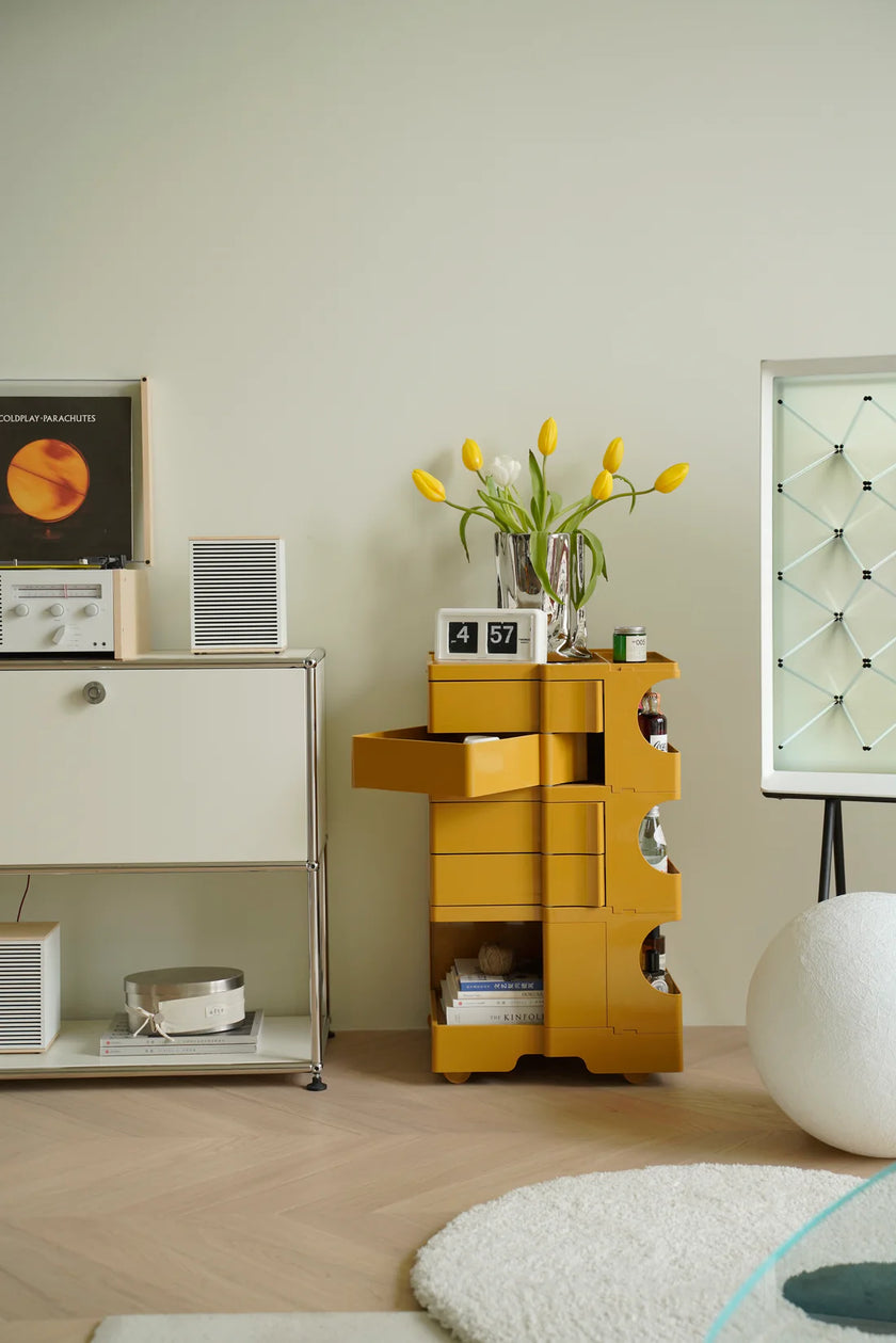 Yellow storage rolling cart with books and flowers in a room with a white cabinet and a plant.