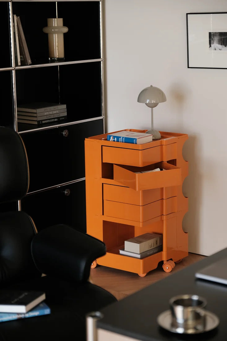 An orange modular rolling storage cabinet with drawers and open shelves, styled in a modern office setting next to a black chair and bookshelf.