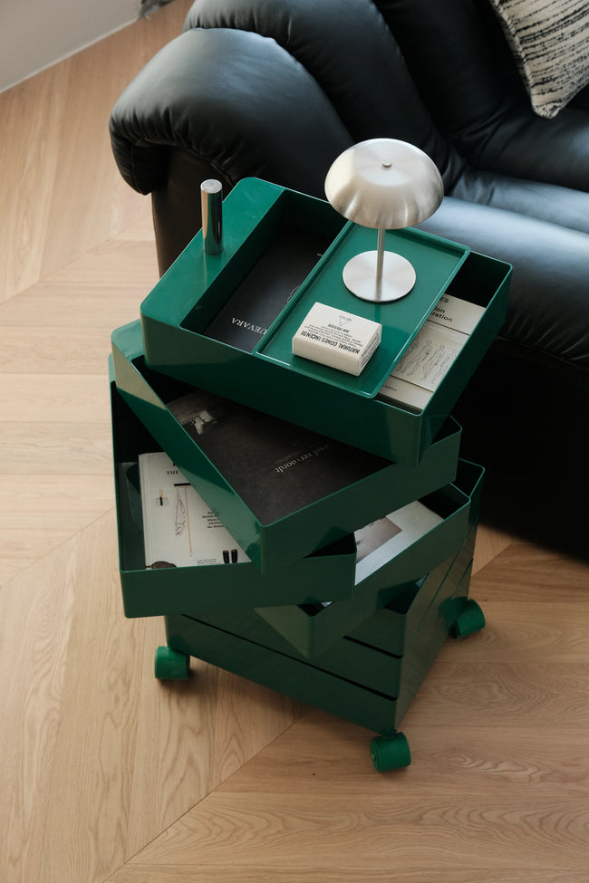 A dark green rolling storage cart with multiple drawers and trays, styled with a white table lamp and books in a modern living room setting with a black leather sofa and light wood herringbone floor.