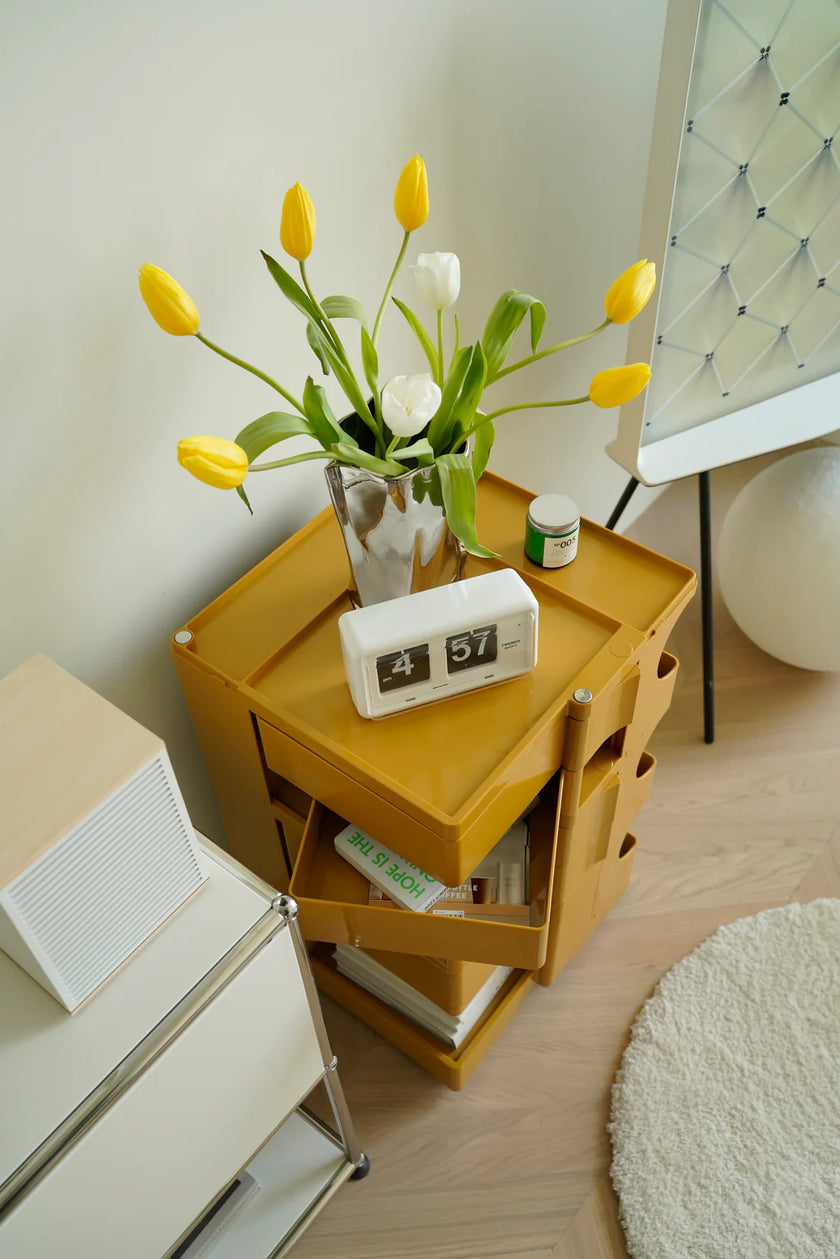Yellow storage cart side table with a vase of yellow and white flowers, clock, and books.