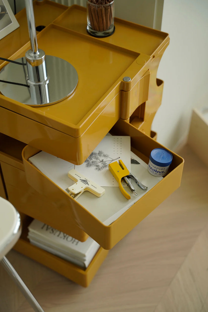 Yellow 3 tier storage rolling cart with open drawer containing stationery items on a wooden surface.
