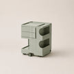 A minimalist, modern rolling storage cart in a muted matcha green color, featuring two drawers on the left, two open shelves with circular cutouts on the right, and small wheels at the base, set against a solid light beige background.