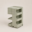 A light matcha green plastic rolling organizer cart with two drawers on the top left, three open shelves with curved cutouts, and small caster wheels, shown on a plain beige studio background.