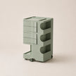 A modern three-tier rolling storage cart in a muted sage green color, featuring drawers on the top two levels and open shelves on the bottom, set against a solid light beige background.
