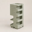 A light sage green rolling storage cart with multiple tiers, open shelves, and a stack of drawer fronts on the left side, set against a solid light beige background.