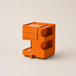 An orange ABS plastic storage cart with two drawers on the left, two open shelves on the right, and small wheels at the base, photographed on a light beige studio background.