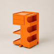 Orange rolling storage cart with two drawers, two open shelves, and open side compartments, photographed on a plain light gray studio background.
