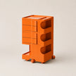 An orange storage cart with multiple drawers, open shelves, and small caster wheels, photographed on a clean light beige studio background.