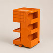 An orange rolling storage cart with multiple tiers, including a stack of drawers on the upper left, open shelves, and caster wheels, photographed on a neutral light gray background.