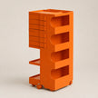 An orange modular rolling storage cart with multiple drawers, open shelves, and wheels, photographed on a neutral light beige background.