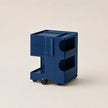 A steel blue ABS plastic storage cart with two drawers, open shelves, and small black wheels, photographed on a clean, solid, warm off-white background.