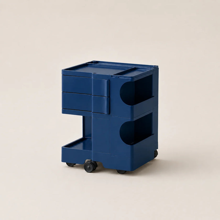 A steel blue ABS plastic storage cart with two drawers, open shelves, and small black wheels, photographed on a clean, solid, warm off-white background.