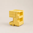A small tulip yellow ABS plastic storage cart with three drawers on the left, two open shelves on the right, and caster wheels at the base, photographed on a plain light beige studio background.