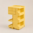 A bright yellow rolling storage cart with two drawers on the left, open shelves in the middle, and a top platform, shown on a plain light beige studio background.