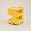 A tulip yellow utility cart with three drawers on the left, three open storage compartments on the front, and a lower tray with caster wheels, shown on a neutral light beige background.