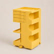 A bright yellow plastic rolling storage cart with three drawers on the top left, three open shelves, and caster wheels, shown on a neutral light beige background.