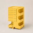 A bright yellow storage organizer with multiple drawers, open shelves, and a wheeled base, photographed on a clean light beige studio background.