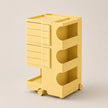 A yellow ABS rolling storage cart with six drawers on the left, three open shelves with curved cutouts on the right, and small caster wheels at the base, shown on a plain light beige studio background.