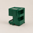 A vintage green plastic rolling storage cart with three drawers on the left, two open shelves on the right, and small caster wheels at the base, photographed on a plain light beige studio background.