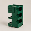 A dark green rolling storage cart with multiple drawers, open shelves, and wheels, photographed on a clean light beige studio background.