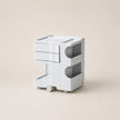A minimalist, white ABS plastic side table or organizer with two drawers, open storage compartments, and small wheels, set against a solid beige background.