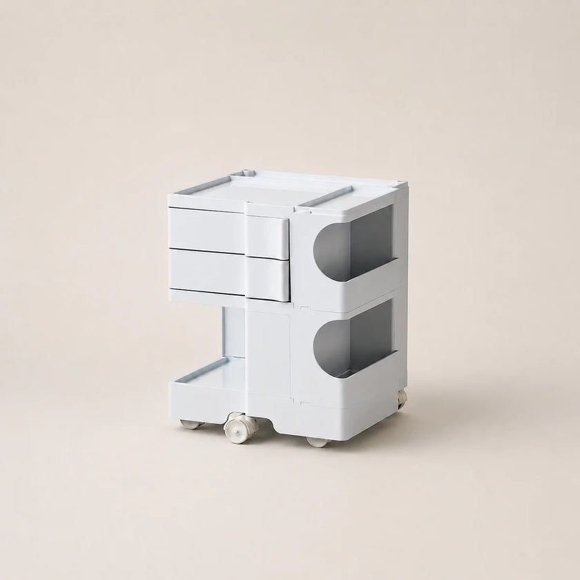 A minimalist, white ABS plastic side table or organizer with two drawers, open storage compartments, and small wheels, set against a solid beige background.